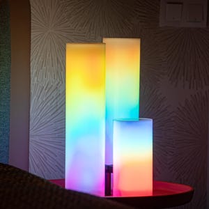 May include: Three rectangular LED lamps in varying heights, each displaying a gradient of colors. The tallest lamp transitions from yellow to blue, while the others feature similar color schemes. The lamps are on a small, round, red table.
