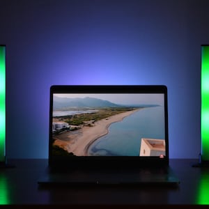 Peut inclure: Un ordinateur portable affichant une scène côtière est placé sur une surface sombre, flanqué de deux lampes LED verticales vertes. L'arrière-plan passe du bleu au violet, créant une ambiance moderne et colorée.