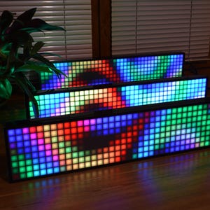 Può includere: Tre pannelli di visualizzazione a LED rettangolari sono impilati, ognuno mostra un motivo vibrante e colorato. I pannelli sono incorniciati in nero ed emettono uno spettro di colori, tra cui rosso, verde, blu e giallo. I display sono su una superficie di legno.