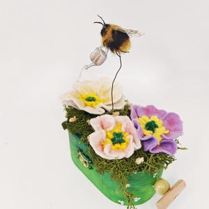 May include: A whimsical, handmade felt flower arrangement in a green container. The arrangement features three colorful felt flowers, moss, and a miniature bumblebee with a watering can. A wooden dowel and a small ball are next to the container.