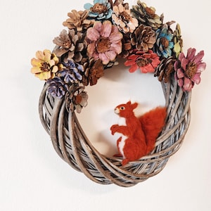 Könnte beinhalten: Ein geflochtener Kranz, der mit bemalten Tannenzapfen und Blumen in Rosa-, Blau- und Gelbtönen verziert ist. Ein kleines, rotes Filz-Eichhörnchen sitzt auf dem Kranz.