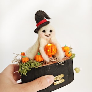 May include: A white felt ghost wearing a black witch hat with a red band holds a jack-o'-lantern. The ghost is sitting in a wooden music box with a black finish and a gold latch. The box is surrounded by small orange felt pumpkins and green moss.