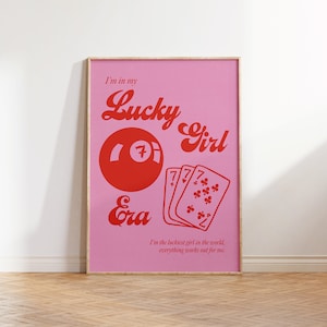 May include: Pink poster with a wooden frame featuring the text "I'm in my Lucky Girl Era." The design includes a red 7-ball, playing cards, and the phrase "I'm the luckiest girl in the world, everything works out for me."