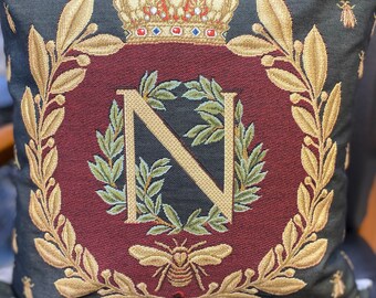 Op de afbeelding: Een zwart en bordeauxrood kussen met een gouden geborduurde krans en een grote letter N in het midden. De krans is gemaakt van bladeren en bessen. Een gouden kroon zit boven de krans. Een bij is geborduurd onder de letter N.