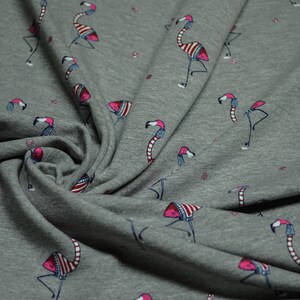 Könnte beinhalten: Grauer Stoff mit einem sich wiederholenden Muster aus rosa Flamingos, die rot-weiß gestreifte Mützen tragen. Die Flamingos sind in verschiedene Richtungen gewandt und haben schwarze Umrisse.