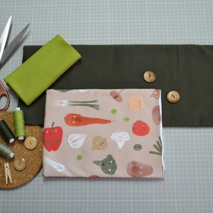 Puede incluir: Una vista plana de los suministros de costura para un proyecto artesanal. Los suministros incluyen dos piezas de tela, una verde y otra con estampado de verduras, carretes de hilo, botones y un par de tijeras.