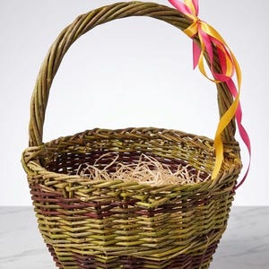 Peut inclure: Un panier en osier tressé, avec une anse, aux tons verts et bruns. Un ruban rose et jaune est noué à l'anse. L'intérieur du panier contient du papier déchiqueté.
