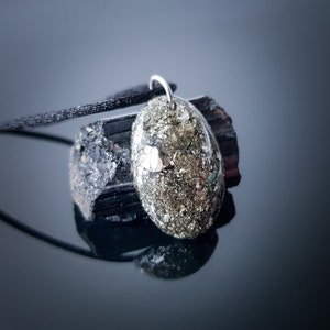 May include: A gold and silver oval pendant with a black cord necklace. The pendant is made of a natural stone with a speckled texture. The pendant is resting on a black stone.