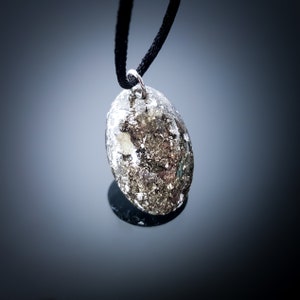 May include: A silver pendant with a black cord necklace. The pendant is a round, flat, oval shape with a silver, metallic, and gold-like speckled pattern.