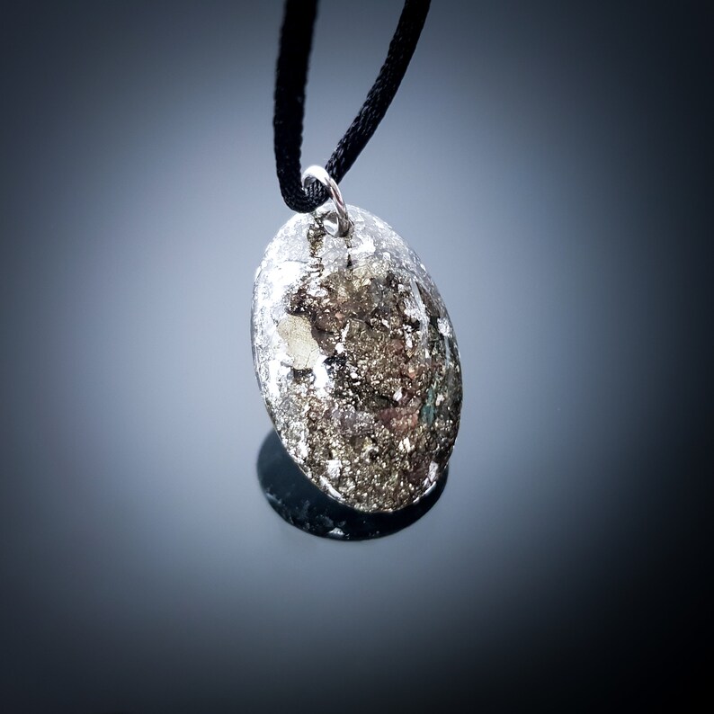 May include: A silver pendant with a black cord necklace. The pendant is a round, flat, oval shape with a silver, metallic, and gold-like speckled pattern.