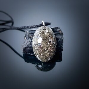 May include: A silver pendant with a gold and black oval-shaped stone. The stone is set in a silver bezel and has a textured surface. The pendant is hanging from a black cord.
