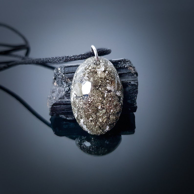 May include: A silver pendant with a gold and black oval-shaped stone. The stone is set in a silver bezel and has a textured surface. The pendant is hanging from a black cord.