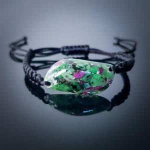 Pode incluir: Um bracelete de cordão preto com um pingente de pedra preciosa verde e rosa. O pingente é feito de resina transparente com pedras preciosas verdes e rosas incrustadas.