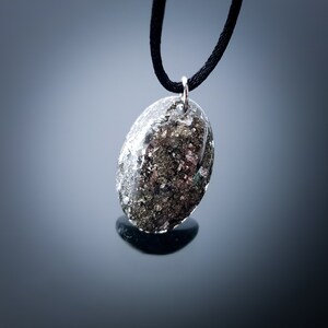 May include: A silver pendant necklace with a black cord. The pendant is oval shaped and filled with a mixture of silver and black flecks.