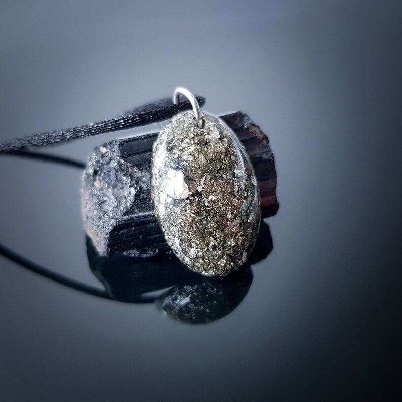 May include: A gold and silver oval pendant with a black cord necklace. The pendant is made of a natural stone with a speckled texture. The pendant is resting on a black stone.