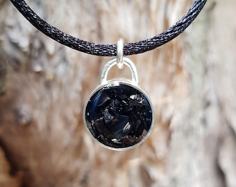 Puede incluir: Un collar con un colgante de piedra negra y una cadena de plata. El colgante es redondo y tiene una superficie texturizada.