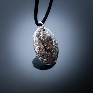 May include: A silver pendant with a black cord. The pendant is oval shaped and has a speckled, gray and silver design.
