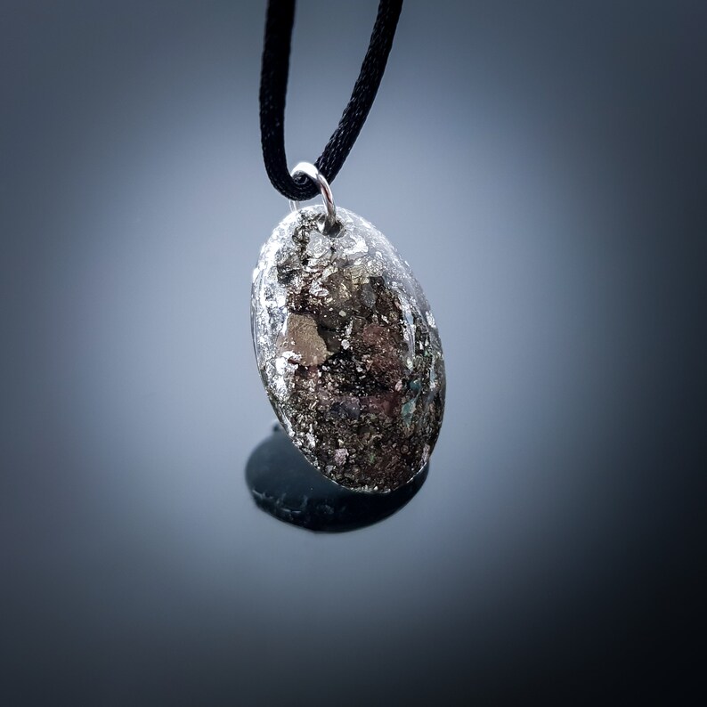 May include: A silver pendant with a black cord. The pendant is oval shaped and has a speckled, gray and silver design.