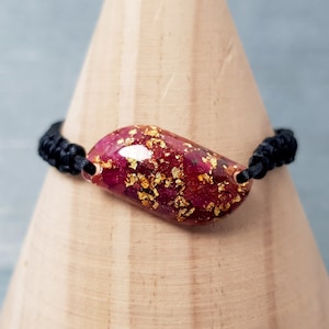 May include: A black cord bracelet with a red and gold resin charm. The charm is shaped like a teardrop and has gold flakes embedded in it.