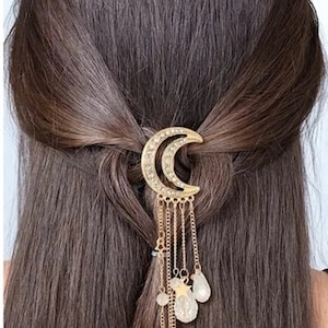 Puede incluir: Pinza para el pelo dorada con un diseño de media luna y cadenas colgantes con estrellas y cuentas transparentes.