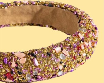 May include: A close-up of a jeweled headband with gold, pink, purple, and clear stones. The headband has a tan interior and is decorated with the text "Ananda: bliss for your hair".