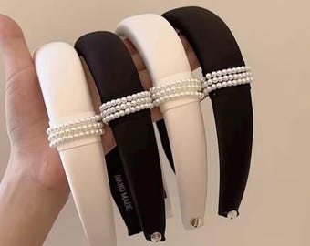 May include: Four headbands in black and white are displayed. Each headband has a thick, padded band with a row of faux pearls. The black headband has the words "HAND MADE" printed on it.