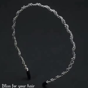 Puede incluir: Una diadema de metal plateado con un diseño retorcido y ornamentado. La diadema presenta un patrón repetitivo de pequeños elementos texturizados y está sobre un fondo oscuro. El texto "Bliss for your hair" está en la parte inferior.