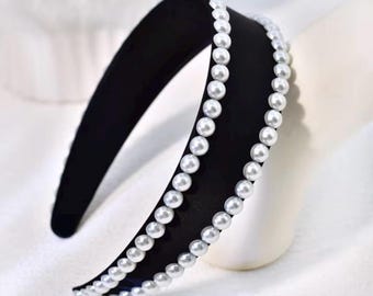May include: A black headband featuring two parallel rows of white, round, lustrous faux pearls. The headband has a smooth, matte finish and a curved, wide structure that arches to form a semi-circle shape, showcasing a clean, uniform design.