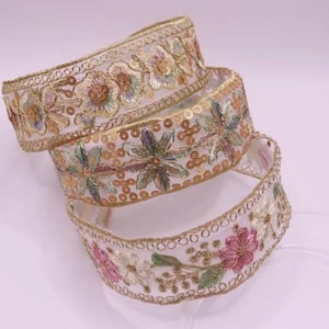 Pode incluir: Três rolos de acabamento floral decorativo. O acabamento é branco com flores bordadas em tons de rosa, verde e dourado. As bordas são adornadas com fio de ouro e lantejoulas. O acabamento é provavelmente usado para costura, artesanato ou embelezamento.
