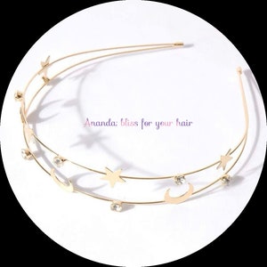 Puede incluir: Diadema dorada con una doble fila de medias lunas y estrellas, adornada con brillantes transparentes. "Ananda bliss for your hair" está escrito en rosa en la diadema.
