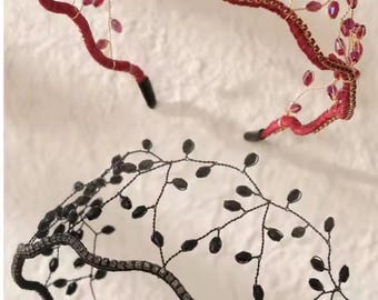 May include: Two headbands featuring a wavy base wrapped in fabric, topped with a delicate wire structure adorned with small, oval-shaped beads. One headband is red with matching red beads, while the other is black with matching black beads.