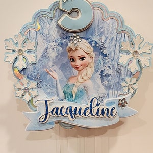 May include: A light blue and white cake topper featuring Elsa from Frozen. The topper has a number 5, snowflakes, and the name "Jacqueline" in a banner. The design includes glitter and rhinestones, perfect for a birthday celebration.