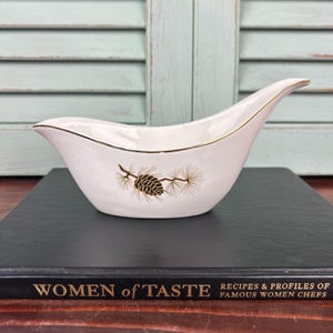 May include: White ceramic gravy boat with a gold rim and a pine cone design. The boat is sitting on a black book with the title "Women of Taste" on the cover.