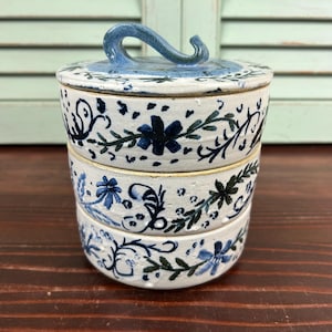 May include: A stackable ceramic container with a lid. The container is white with blue floral and vine designs. The lid has a curved handle. The container is on a wooden surface.