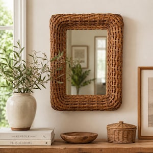 May include: A rectangular wall mirror featuring a thick, woven brown rattan frame. The mirror is mounted on a wall above a wooden surface that holds a cream-colored ceramic vase with green foliage, a small wooden bowl, and a lidded rattan basket.