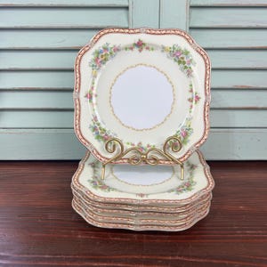May include: A stack of square, cream-colored plates with a floral and gold rim design. The plates are displayed on a gold-colored metal plate stand. The plates have a scalloped edge and a floral pattern with pink, yellow, and green accents.