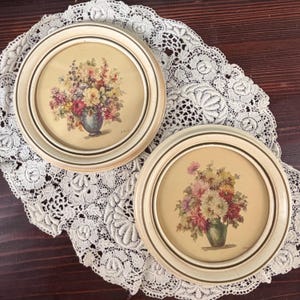 May include: Two circular wall frames with cream-colored borders, each containing a botanical print of a floral bouquet in a vase. The prints feature vibrant flowers in shades of red, yellow, pink, and purple, set against a light beige background.