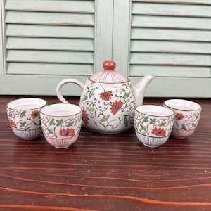 May include: A white ceramic tea set with a teapot and four teacups. The teapot has a red lid and is decorated with red flowers and green leaves. The teacups have a similar floral design with a blue stripe at the base.