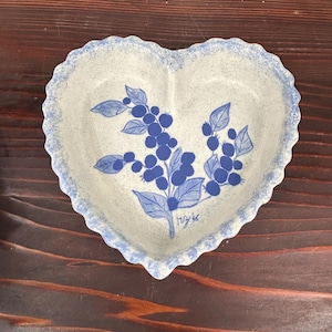 Puede incluir: Plato de cerámica en forma de corazón con un fondo moteado azul claro y blanco. El plato presenta un diseño floral azul con bayas y hojas. El borde tiene un borde festoneado con un borde azul. El plato está sobre una superficie de madera.