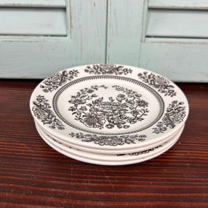 May include: A stack of three white ceramic plates with a black floral design. The plates have a detailed border and a central floral pattern. The plates are stacked on top of each other.