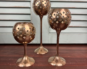 Puede incluir: Tres candelabros de latón con un diseño decorativo calado. Los candelabros tienen forma de cuenco redondo con un tallo largo y una base ancha. Los candelabros están sobre una superficie de madera marrón oscuro, con un fondo de contraventanas azul claro y blanco.