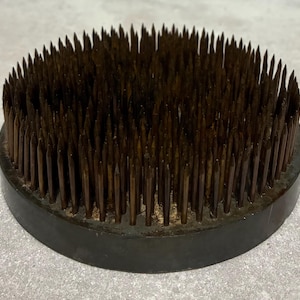 May include: A round, dark brown flower frog with numerous sharp, metal pins. The pins are densely packed and protrude from a circular base. This floral arranging tool is designed to hold flower stems in place.