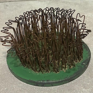 May include: A vintage flower frog, featuring a round, green base with a dark rim. The frog has numerous brown wire stems, creating a dense, textured surface. This floral arranging tool is designed to hold flower stems in place.