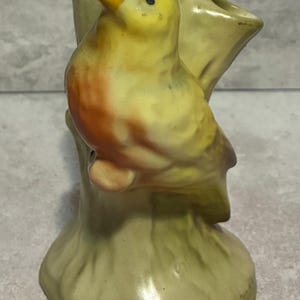 May include: A ceramic bird figurine perched on a tree trunk-shaped vase. The bird has yellow, orange, and blue coloring. The vase is a light green color and has a smooth, glossy finish. The bird is facing left.
