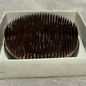 May include: A brown flower frog, also known as a kenzan, with numerous sharp metal pins. The floral arranging tool is set in a black base and presented in a white cardboard box. This tool is used to hold flower stems in place.