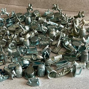 May include: A collection of silver-colored Monopoly game tokens. The tokens include a race car, dog, thimble, iron, shoe, top hat, ship, cannon, and other classic game pieces. The pieces are scattered on a neutral surface.