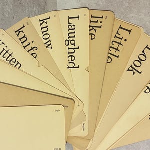 May include: A fan-shaped collection of light tan rectangular educational flashcards featuring black printed words including Jingle, Jump, Just, Kitten, knife, know, Laughed, like, Little, Look, Make, and Makes, arranged in a layered, overlapping circular pattern.