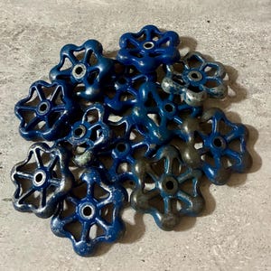May include: A collection of vintage blue water valve handles. The handles have a flower-like design with a central hole and are made of metal. Some handles show signs of wear, with the blue paint partially worn away, revealing the metal underneath.