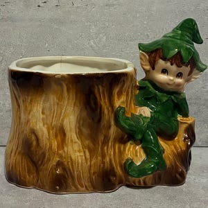 May include: A ceramic planter shaped like a tree stump with a smiling elf figure. The planter is brown with a white interior. The elf is wearing a green outfit and hat. The planter is a decorative item.