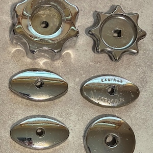 May include: A collection of shiny, chrome-plated hardware components. The assortment includes two star-shaped knobs and four oval-shaped pieces, some with the text "EASTMAN SPEEDLINK" engraved. The pieces are arranged on a neutral-toned surface.